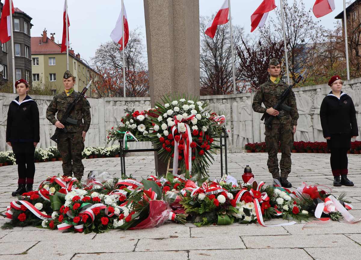 Pod pomnikiem stoją żołnierze i harcerze, widoczne złożone kwiaty i flagi.