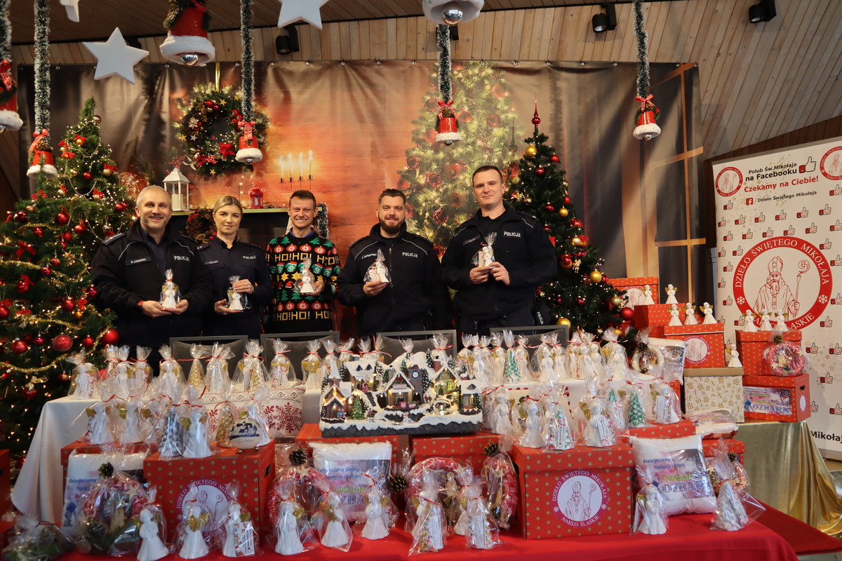 Zdjęcie przedstawia policjantów i organizatora akcji Dzieło Świętego Mikołaja, którzy stoją wśród prezentów i ustrojonych choinek.