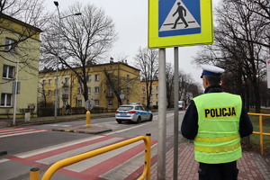 Policjant ruchu drogowego stoi przy przejściu dla pieszych widoczny też zaparkowany radiowóz