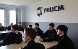 Uczniowie siedzą na auli w komendzie, nad nimi, na ścianie napis &quot;Policja&quot;.