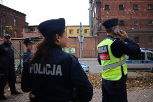 Policjant w kamizelce z napisem &quot;Dowódca akcji&quot; rozmawia przez telefon, obok stoi policjantka i funkcjonariusze służby więziennej.