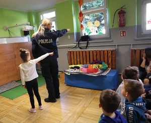 Policjantka i dziewczynka pokazują przedszkolakom, w jaki sposób rozglądamy się przed przejściem przez jezdnię.