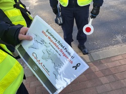 Na zdjęciu widoczny policjant, który trzyma plakat z napisem &quot;Światowy Dzień Pamięci Ofiar Wypadków Drogowych&quot;, za nim policjant trzyma tarcze do zatrzymywania pojazdów.