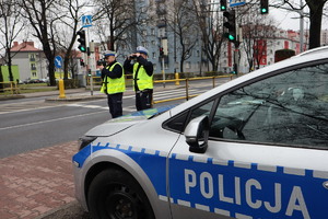Na zdjęciu widoczny radiowóz i dwaj policjanci ruchu drogowego stoją przy przejściu, jeden mierzy prędkość, drugi patrzy przez lornetkę.