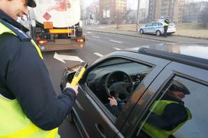 Zdjęcie przedstawia policjanta ruchu drogowego, który sprawdza trzeźwość kierowcy, widać również stojącego przy radiowozie drugiego policjanta i jadące samochody.