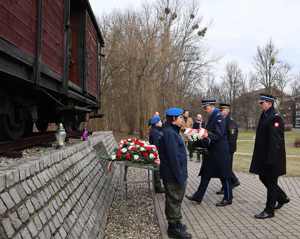 Na zdjęciu widzimy delegację kierownictwa jednostek Policji, Straży Miejskiej i Straży Pożarnej, jak idą z kwiatami w stronę pomnika.