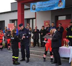 Na zdjęciu widzimy, jak Zastępca Komendanta Miejskiego Policji w Bytomiu wręcza puchar uczestniczce Śląskich Mistrzostw w Pierwszej Pomocy przed budynkiem, obserwują to inni uczestnicy.