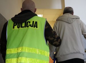 Zdjęcie przedstawia policjanta w kamizelce z napisem Policja, który prowadzi zatrzymanego.