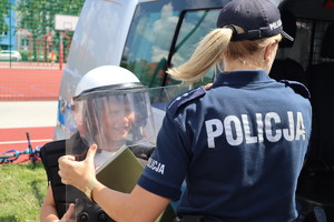Policjantka ubierająca chłopcu policyjny sprzęt, to jest kamizelkę, tarczę i kask.