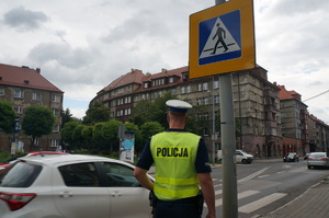 Na zdjęciu widzimy policjanta, który sto przy oznakowanym przejściu dla pieszych i przejeżdżający samochód.