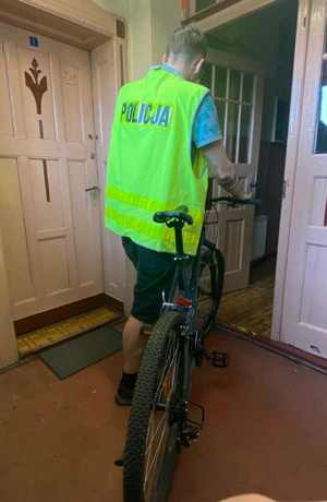 Na zdjęciu widzimy policjanta z odblaskową kamizelką z napisem Policja, który wprowadza rower do pomieszczenia.