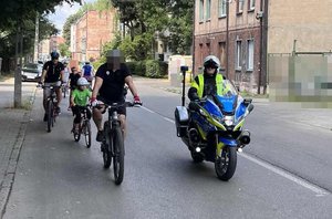 Na zdjęciu widzimy policjanta, który jedzie na motocyklu, a za nim jadą rowerzyści.