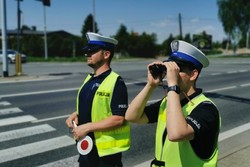 Zdjęcie przedstawia dwóch policjantów ruchu drogowego, którzy stoją przy drodze, a jeden z nich patrzy przez lornetkę.