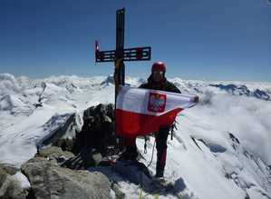 Zdjęcie przedstawia mężczyznę trzymającego polską flagę na szczycie góry przy krzyżu.