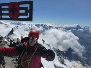 Zdjęcie przedstawia alpinistę, w oddali panorama szczytów w chmurach.