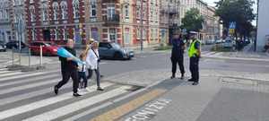 policjant i strażniczka miejska stojący przy skrzyżowaniu, rodzice z dzieckiem przechodzący przez ulicę przejściem dla pieszych