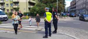 dorośli i dzieci przechodzący przez ulicę na pasach, policjant i strażniczka miejska stojący przy skrzyżowaniu