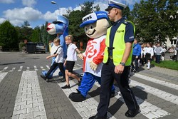 Zdjęcie przedstawia umundurowanego policjanta, maskotki oraz dzieci przechodzące przez przejście dla pieszych.