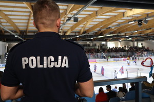 Na zdjęciu widzimy policjanta, który patrzy w stronę tafli lodowiska, na której rozgrywany jest mecz.