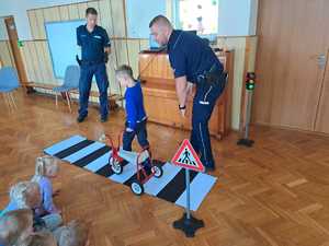 Zdjęcie przedstawia policjantów w przedszkolu, którzy pokazują chłopcu, jak prawidłowo przechodzić przez przejście dla pieszych.