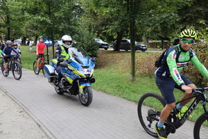 Zdjęcie przedstawia rowerzystę w parku, za nim jadący policyjny motocykl i rowerzystów.