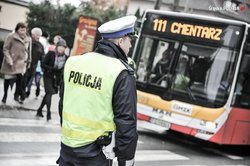 Na zdjęciu widzimy policjanta przy przejściu dla pieszych, przechodzących przez przejście pieszych i autobus.