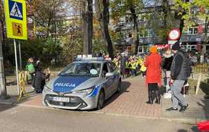 Zdjęcie przedstawia wyjeżdżający z terenu szkoły radiowóz, a za nim  biegnący uczniowie.