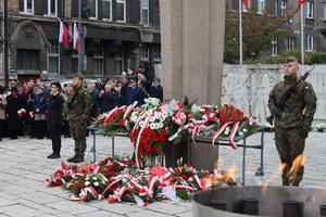 Na zdjęciu widzimy pomnik z kwiatami, w tle przedstawiciele służb mundurowych, władz miasta i zebrani uczestnicy obchodów.