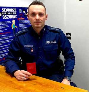 Policjant siedzi przy biurku i trzyma legitymację Zasłużonego Honorowego Dawcy Krwi.