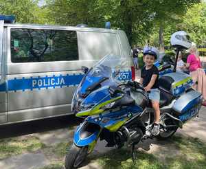 Na zdjęciu radiowóz policyjny, przed nim chłopiec siedzący na motocyklu służbowym, na głowie czapka policyjna.