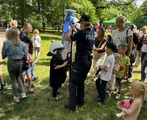 Na zdjęciu policjantka, pomaga kilkuletniej dziewczynce założyć sprzęt policyjny, wokół niej osoby