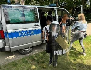 Na zdjęciu radiowóz policyjny, obok niego osoby, przed nim chłopiec ubrany w sprzęt policyjny, kamizelkę, tarczę, kask, trzyma w dłoni pistolet.