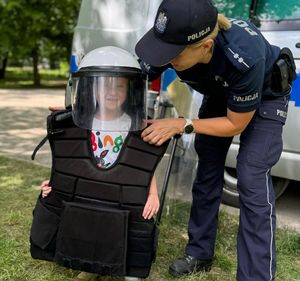 Na zdjęciu kilkuletnie dziecko, ma na sobie sprzęt służbowy czarną kamizelkę, biały kask, w ręku trzyma tarcze. Obok pochylona nad dzieckiem policjantka.