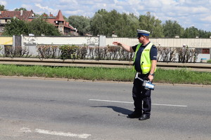 Na zdjęciu policjant w żółtej kamizelce z uniesioną ręką, w drugiej dłoni trzyma urządzenie do pomiaru prędkości.