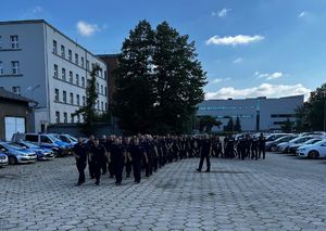 Na zdjęciu idący czwórkami z zwartym szyku policjanci w granatowych mundurach, obok nich jeden funkcjonariusz. Idą po wybrukowanym placu, obok widoczny budynek i zaparkowane dookoła placu radiowozy i pojazdy.