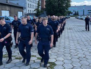 Na zdjęciu idący czwórkami z zwartym szyku policjanci w granatowych mundurach, obok nich jeden funkcjonariusz. Idą po wybrukowanym placu, obok widoczny budynek i zaparkowane dookoła placu radiowozy i pojazdy.