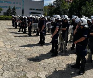 Na zdjęciu stojący na placu policjanci, ubrani w czarne kamizelki taktyczne, białe kaski, część z nich trzymają tarcze.