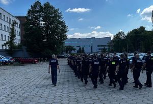 Na zdjęciu idący czwórkami z zwartym szyku policjanci w granatowych mundurach, obok nich jeden funkcjonariusz. Idą po wybrukowanym placu, obok widoczny budynek i zaparkowane dookoła placu radiowozy i pojazdy.