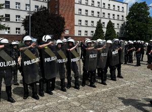 Na zdjęciu stojący na placu policjanci, ubrani w czarne kamizelki taktyczne, białe kaski, trzymają przezroczyste tarcze z napisem Policja.