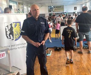 Na zdjęciu policjant w mundurze, za nim osoby zgromadzone w dużej sali, cześć z nich znajduje się na niebieskich matach rozłożonych na środku pomieszczenia.