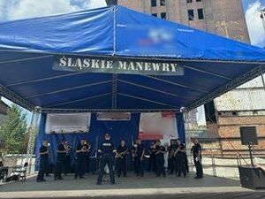 Na zdjęciu policjanci z instrumentami znajdujący się na scenie, w trakcie występu, nad nimi zadaszenie niebieskiego koloru, na nim napis Śląskie Manewry.