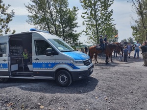 Na zdjęciu radiowóz policyjny, obok dwa konie z policjantami na grzbietach.