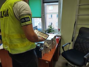 Na zdęciu policjant w żółtej kamizelce i opasce na ramieniu z napisem policja, obok leżące na biurku paczki papierosów.