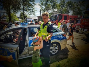 Na zdjęciu policjant przekazujący swoją czapkę kilkuletniemu chłopcu, dookoła zgromadzone osoby, radiowóz policyjny, wozy strażackie.