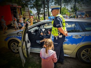 Na zdjęciu policjant, obok niego kilkuletnia dziewczynka, przy nich siedzący w radiowozie policyjnym kilkuletni chłopiec. Dookoła zgromadzone osoby.