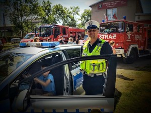 Na zdjęciu policjant przekazujący obok niego radiowóz policyjny, w którym za kierownicą znajduje się chłopiec, dookoła zgromadzone osoby, wozy strażackie.