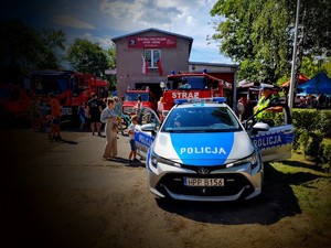 Na zdjęciu radiowóz policyjny, przy nim policjant, za nim pojazdy i budynek straży pożarnej, dookoła przemieszczające się osoby.