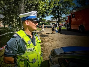 Na zdjęciu policjant ruchu drogowego spoglądający na uczestników, obok widoczny wóz strażacki.