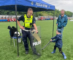 Na zdjęciu stoisko bytomskich mundurowych, przy nim policjant, który trzyma policyjną tarczę, kilkuletni chłopiec uderza w nią pałką służbową.