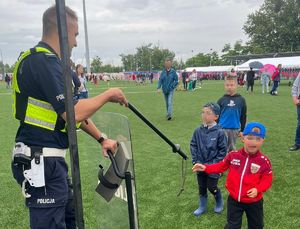 Na zdjęciu policjant i troje chłopców, policjant wręcza im pałkę policyjną.